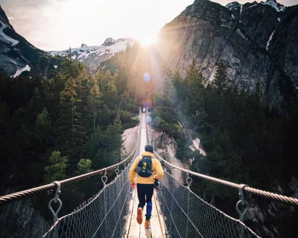 Ein Mann läuft eine Hängebrücke entlang, auf einen Berg zu, hinter dem gerade die Sonne untergeht.