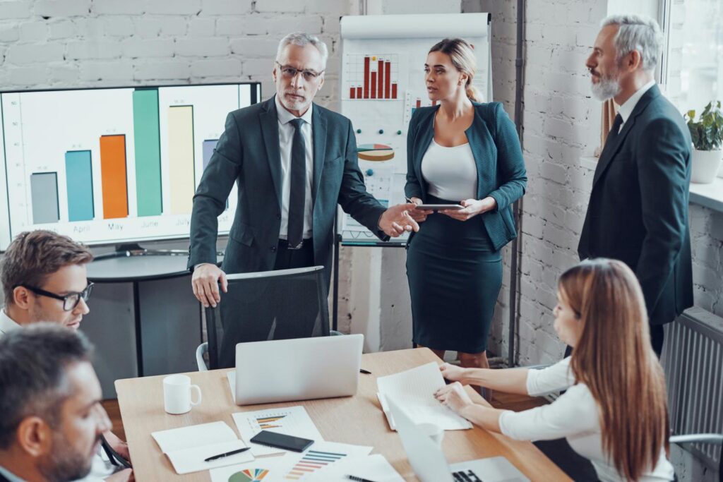 Eine Gruppe von Menschen in Business-Kleidung steht und sitzt um einen Tisch in einem Meetingraum herum. Im Hintergrund ist ein großer Bildschirm und ein Whiteboard zu sehen, die beide mit unterschiedlichen Balkendiagrammen versehen sind.