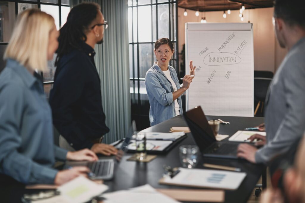 Eine Person steht neben einem Whiteboard mit Aufzeichnungen und hält einen Vortrag vor einigen stehenden Personen in einem Büro.