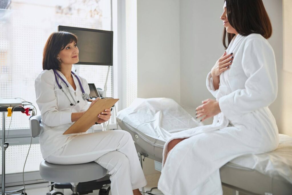 A sympathetic doctor sits on a stool in an examination room and takes notes. In front of her, a patient in a white bathrobe sits on an examination couch and holds her right hand to her chest with her eyes closed.