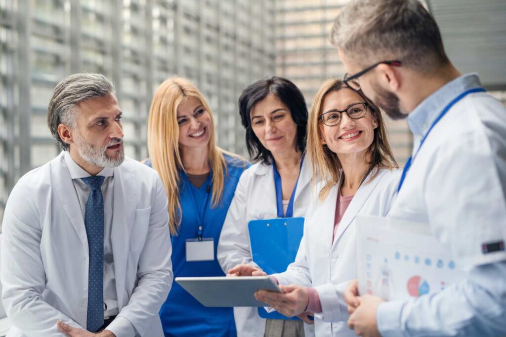 A group of male and female doctors and nurses appear to be standing in a light-flooded hospital corridor, discussing matters. A woman holds a tablet in her hand and points at something with a smile. 