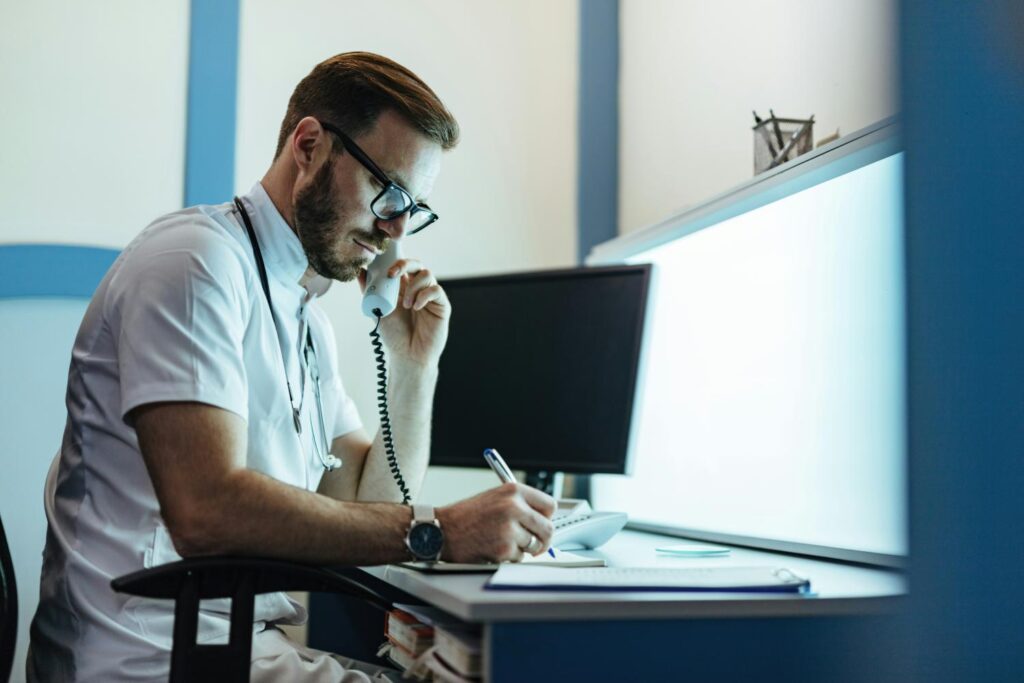 A doctor sits at his desk on the phone and takes notes. In the background is a black computer screen, in front of it is a light screen for x-rays and the like.