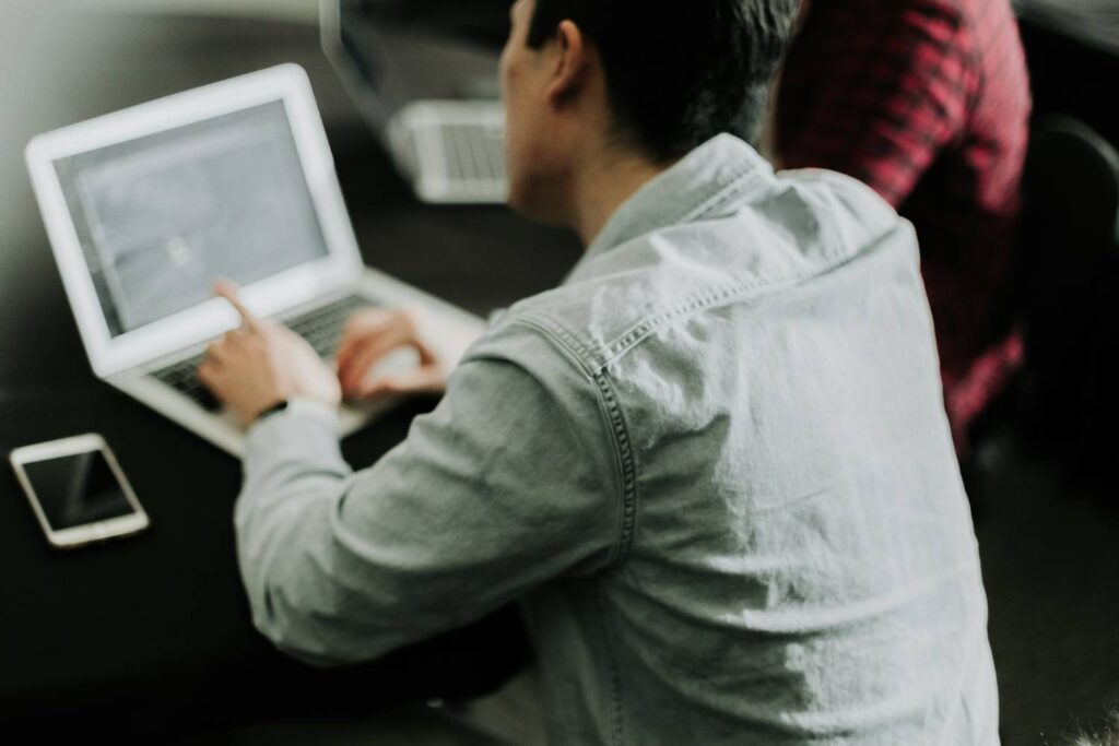 A dark-haired man in a light gray denim shirt is sitting at a black table in front of an open silver laptop whose screen he is pointing at. To his left is a smartphone. To his right is another person in a red plaid shirt sitting in front of an open laptop.