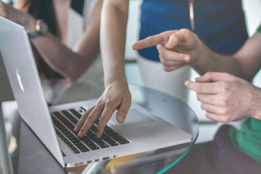 Man points with outstretched index finger at silver laptop screen on which a woman is typing something.