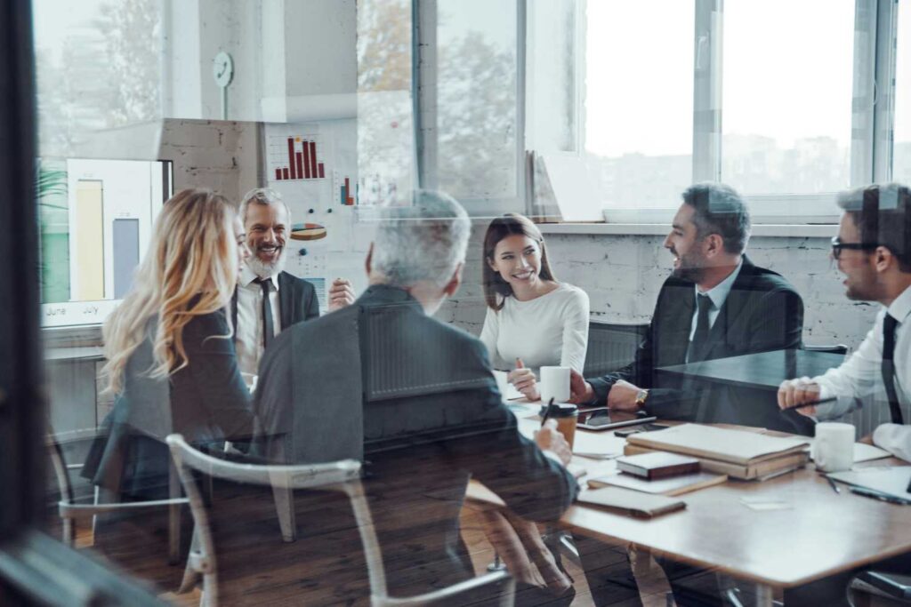 Das Bild zeigt eine Gruppe von Geschäftsleuten, die um einen Tisch sitzen und sich unterhalten. Die Personen scheinen professionell gekleidet zu sein und es ist ein eher formeller Kontext. Der Hintergrund ist ein Büro mit Fenstern und weißen Wänden.