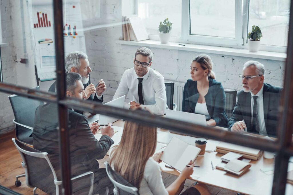 The picture shows a group of business people sitting at a table in a conference room and chatting. They are sitting around a table and appear to be discussing business. You can see laptops on the table and papers, indicating that they are engrossed in a meeting. The atmosphere is professional and serious.