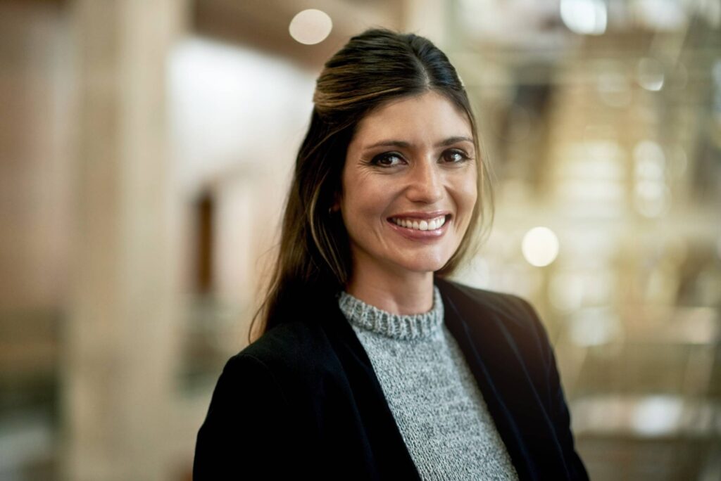 Portrait of a dark-haired woman smiling friendly into the camera. She stands in front of a blurred background.