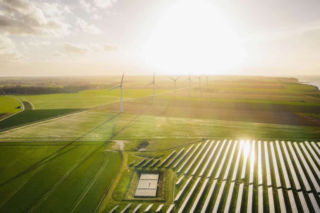 Das Bild zeigt eine Luftaufnahme einer ländlichen Landschaft, in der sich Windkraftanlagen und Solarfelder befinden. Die Sonne scheint von rechts und beleuchtet die Solarpaneele, die in langen Reihen angeordnet sind. Die Windkraftanlagen stehen in der Ferne auf einem Feld. Die Landschaft besteht aus grünen Feldern und ist von einem hellen Himmel begrenzt. Das Bild vermittelt einen Eindruck von nachhaltiger Energieerzeugung und friedlicher Natur.