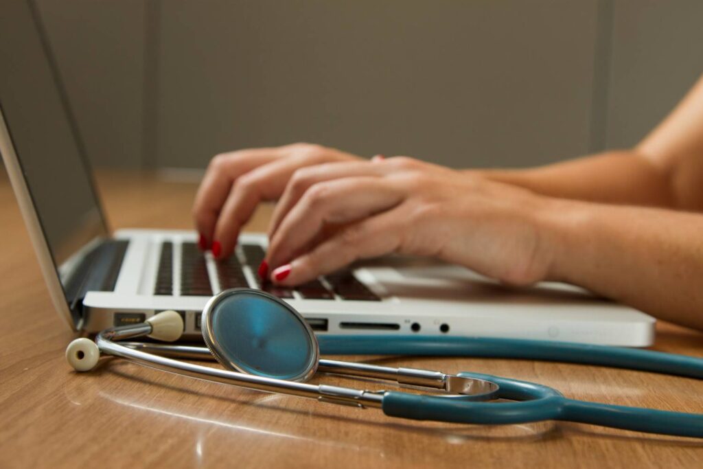 Female hands with red nail polish type on a silver laptop standing on a wooden table.. In the foreground is a stethoscope.