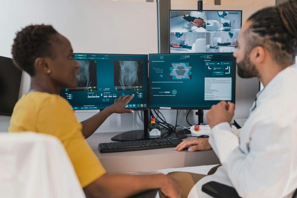 A black woman is talking to a man, also of color, who is wearing a smock and points to a computer screen on which an X-ray image is displayed