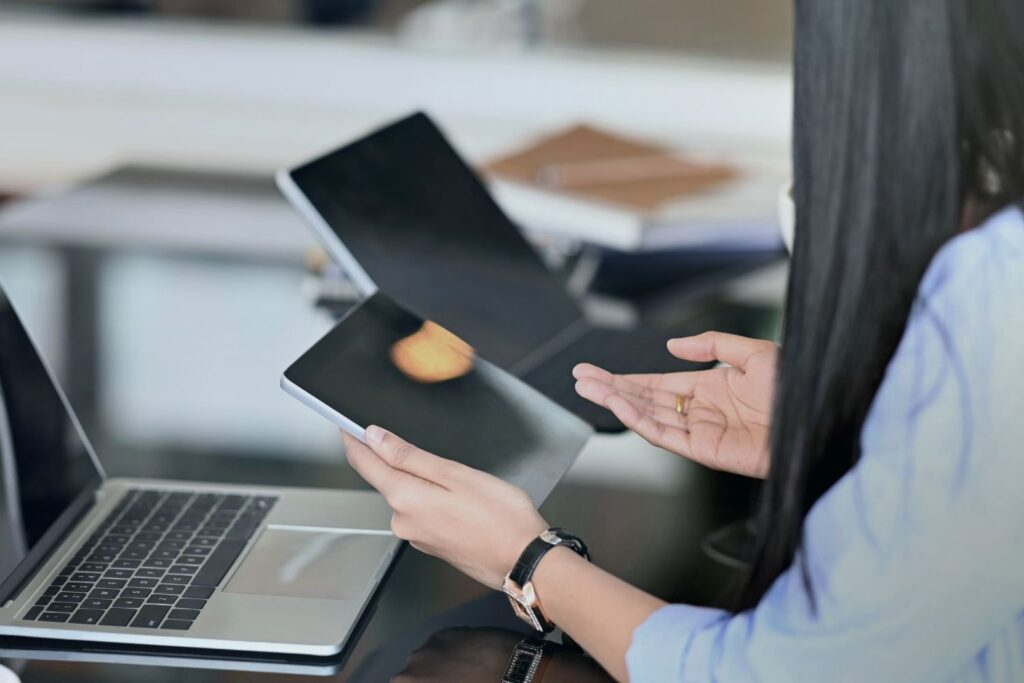 A woman sits at a desk holding a tablet in her left hand while gesticulating with her right hand. There is a laptop on the desk in front of her. The scene suggests a business or professional meeting.