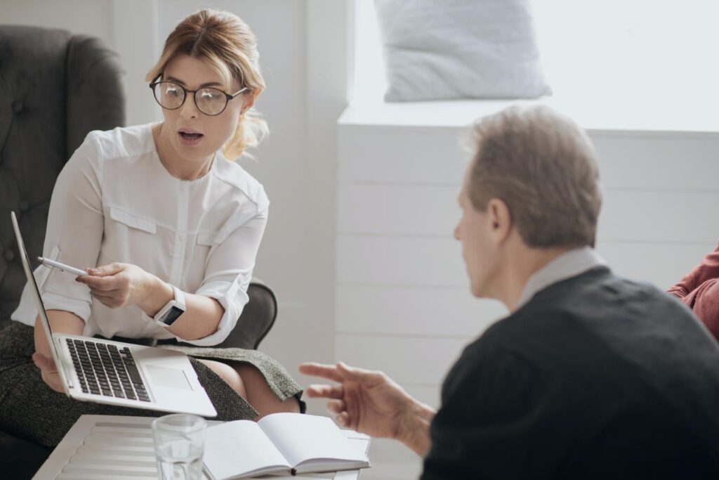 A blonde woman with glasses and a man sit opposite each other. She holds an open, silver laptop in one hand and points to the screen with the other and a pen. The man also stretches out his index finger. Between them are papers and an open book, as well as a glass of water. 