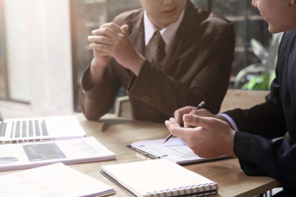 Two people are sitting at a wooden table, one in a brown suit with a shirt and tie, the other is cut and wearing black. They sit facing each other and talk to each other. In front of them are a clipboard, a spiral pad, various other papers and an open silver laptop.