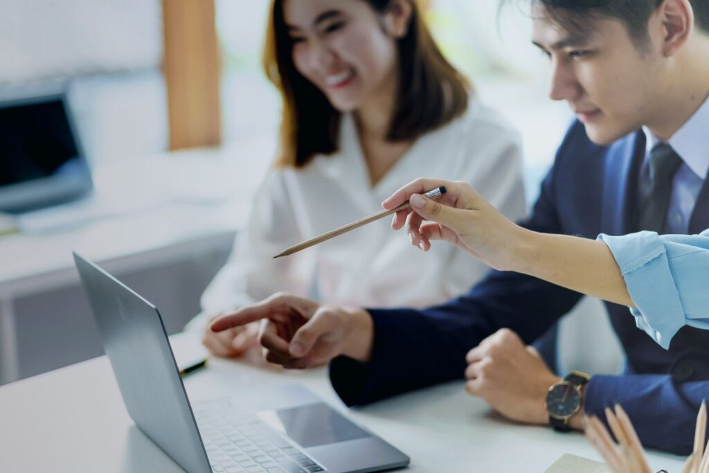 Two women and a man in a suit with a shirt and tie are sitting in front of an open silver laptop on a white table in front of them. The man and the woman, of whom only the hand is visible, point at the screen. The other woman smiles kindly as she also looks at the screen. 