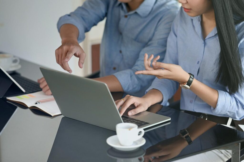 A man and a woman sit at a dark screen, with only their upper bodies visible. In front of them is a silver, open laptop, an open notebook, a pencil, two coffee cups and a tablet. Both point their index fingers at the laptop screen.