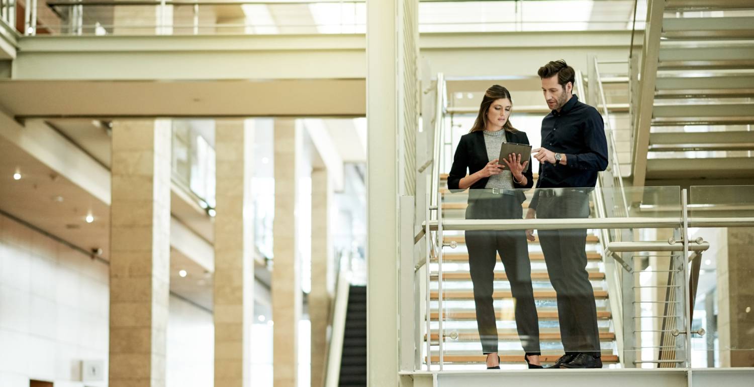 A woman and a man are talking on a staircase about something shown on the woman's tablet. The staircase is in a bright, open-plan office.