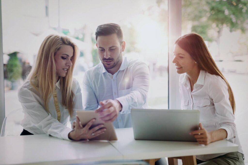 A man and two women are sitting at a white table in front of a large window. The man sits in the middle and, together with the woman on his right, holds a laptop as if he wants to take it from her. All three are looking at this tablet screen. In front of the other woman is an open silver laptop. Overall, the picture is very bright, partly due to the white shirts worn by the three people.