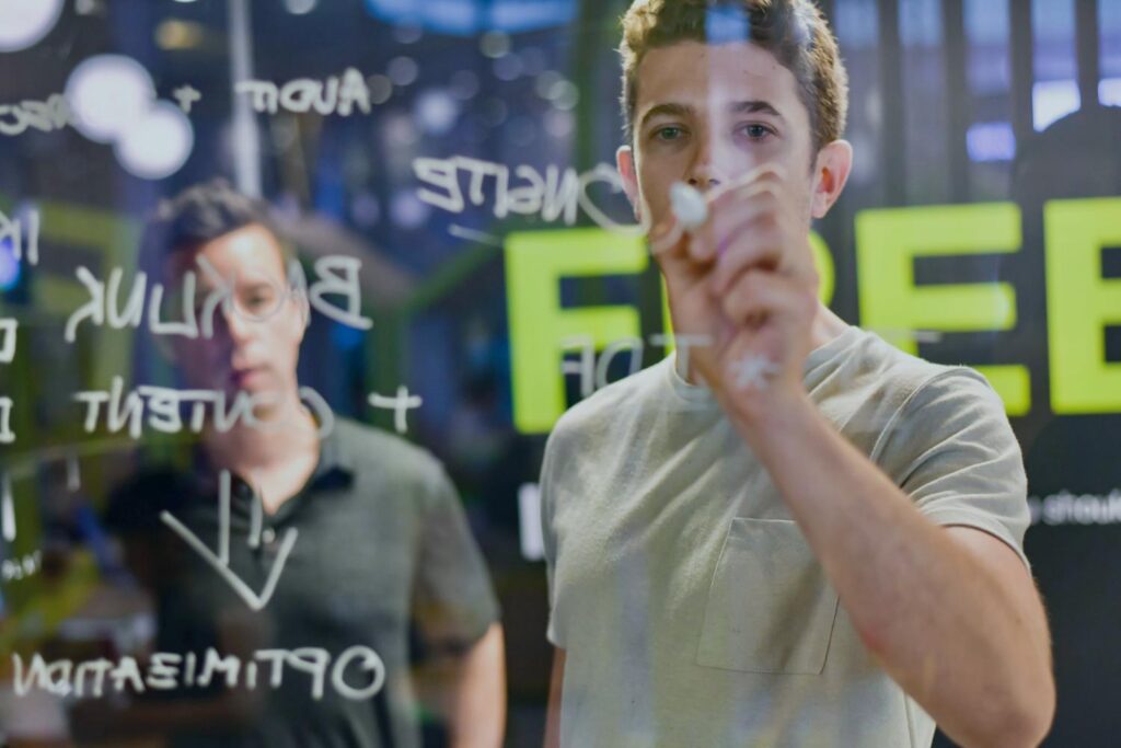 Two men stand in front of a transparent board on which various words and diagrams are written with a white marker. The man in the foreground is writing something, while the man in the background is looking at the blackboard. The scene takes place in a modern, well-lit room.