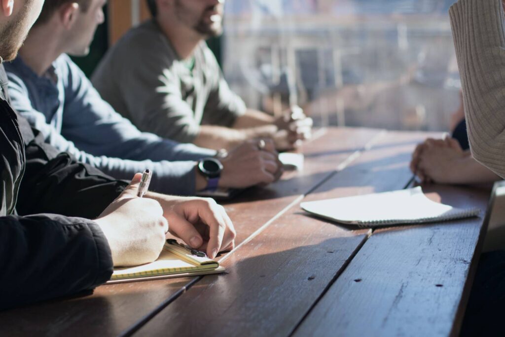 
Several people sit at a wooden table and take part in a meeting. Most have notepads and pens in front of them, while one person wears a wristwatch. The scene is brightly lit by the incoming daylight.