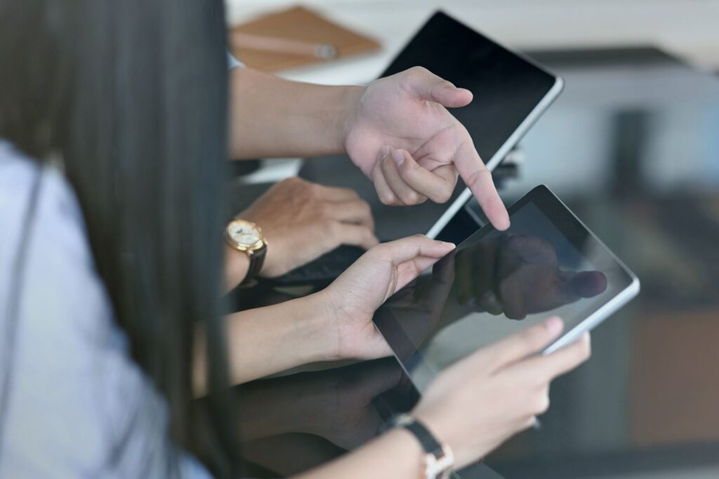 A man and a woman each have a tablet in their hands, which are clearly in the foreground. The man points to the woman's tablet. What is displayed on the screens is not visible to the viewer.