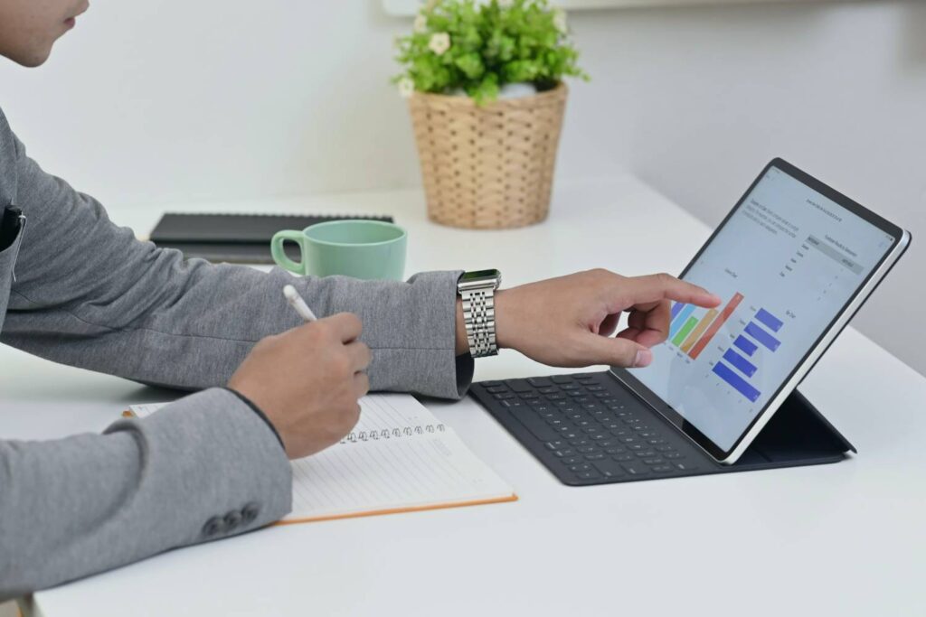 A man in a gray suit sits at a white desk and works on a tablet with a keyboard. Colorful bar charts are displayed on the tablet screen. He holds a pen in one hand and points at the screen with the other. There is an open notebook, a green cup and a plant pot on the table.