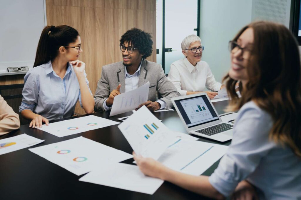 A group of five people sit at a conference table and discuss diagrams and reports. Three of them are women and two are men. A woman and a man in the center of the picture are talking and holding documents in their hands. Another woman is sitting in front of a laptop with diagrams on the screen. Everyone seems focused and committed. There are various documents with colored diagrams on the table.