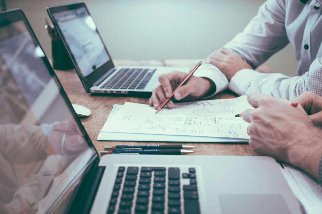 Zwei Personen arbeiten zusammen an einem Schreibtisch, auf dem zwei Laptops und ein Stapel Papier liegen. Beide Personen halten Stifte in der Hand.