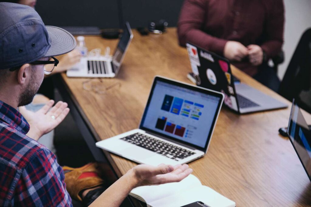 Group work with laptops - different people sit at a wooden table. In the foreground is a man wearing a blue cap and a blue, white and red checked shirt. He has both hands stretched out in front of him, palms facing upwards. 