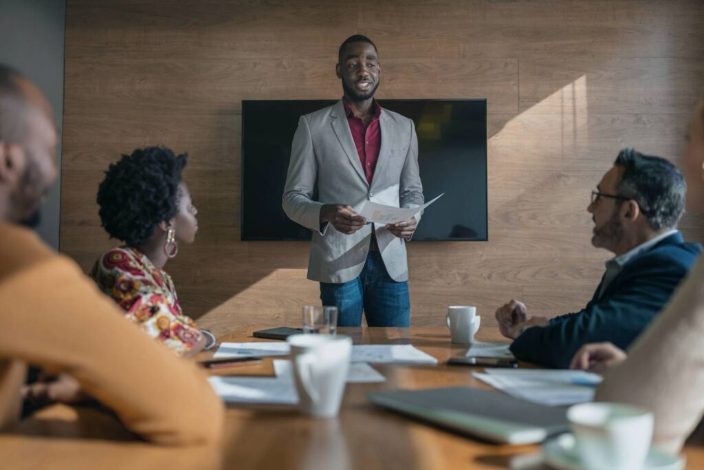 In a conference room with wooden wall paneling, a black man in a burgundy shirt and gray jacket stands in front of a group of people sitting at a wooden table looking at him while he gives what appears to be a lecture. He has a printed paper in his hand. In front of the others on the table are more papers, as well as white coffee cups and drinking glasses.