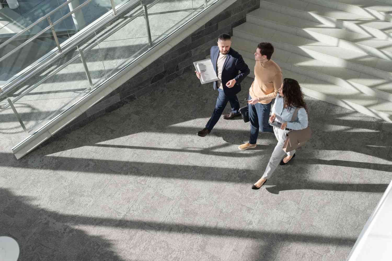 Zwei Männer und eine Frau laufen gemeinsam durch ein modernes, mit Tageslicht hell beleuchtetes Gebäude und unterhalten sich. Die Kamera schaut von oben auf die drei herunter.