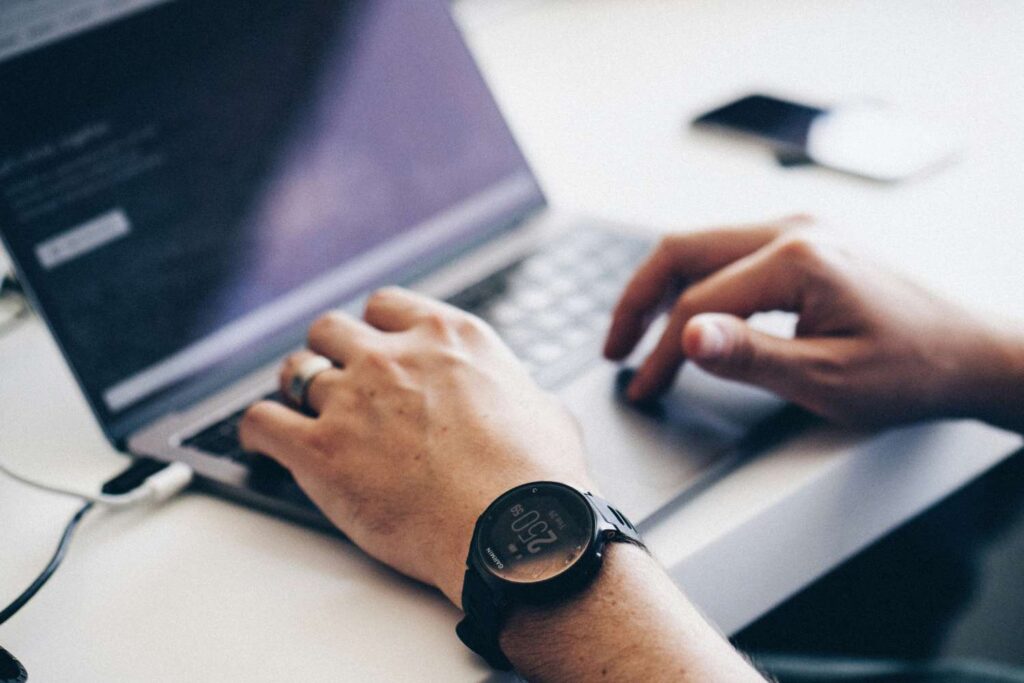 The viewer can recognize male hands working on a laptop standing on a white table. To the right of the laptop is a smartphone. The man is wearing a broad silver ring on his left hand and a black digital watch. 